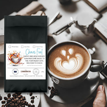A bag of Chaos Fuel – Dark Roast Sumatran Coffee ☕ sits near a latte with heart-shaped foam, spoon, and saucer, surrounded by coffee beans on a wooden table—crafted in small batches and air-roasted for rich flavor.